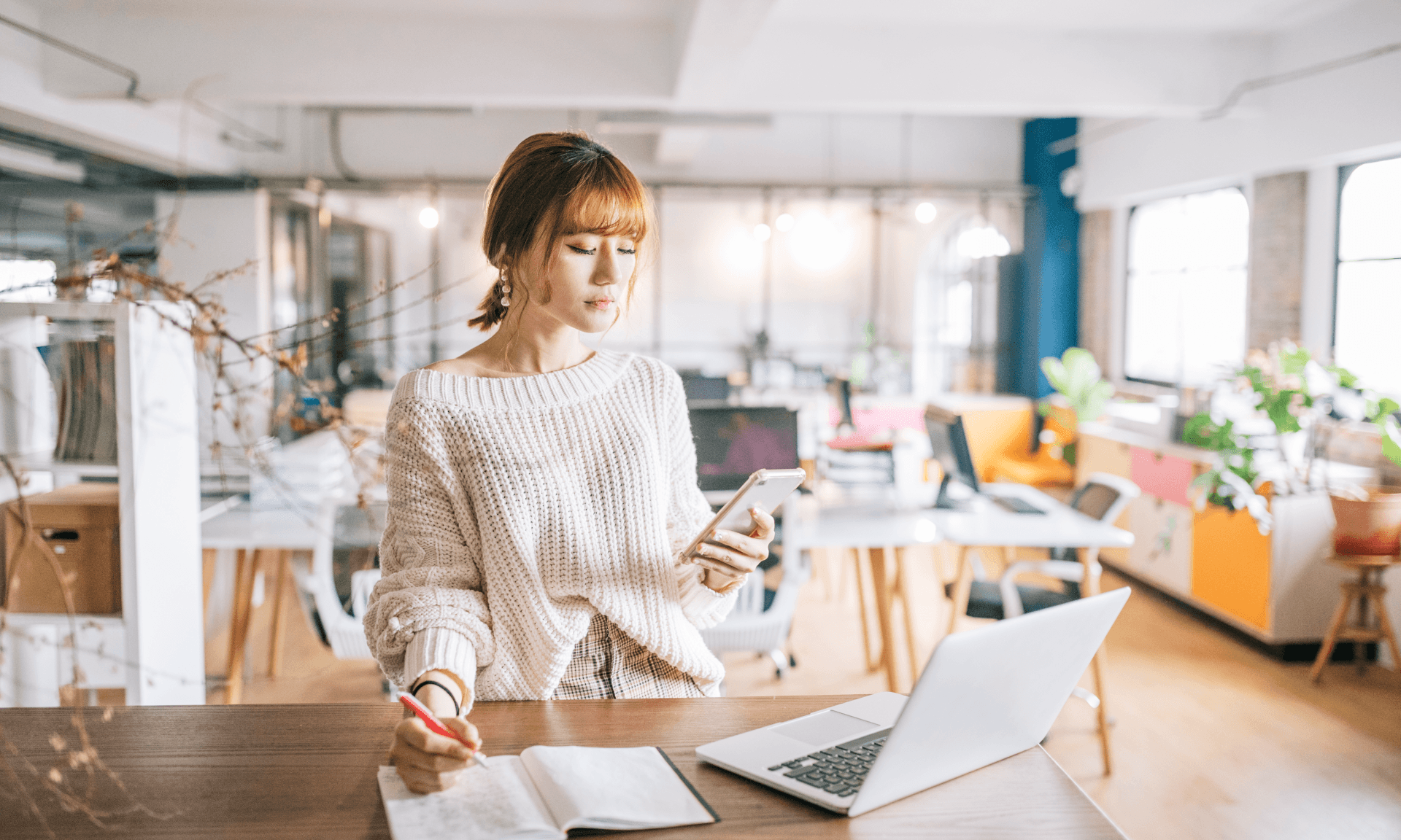 Woman looking at phone while standing in front of computer and datebook